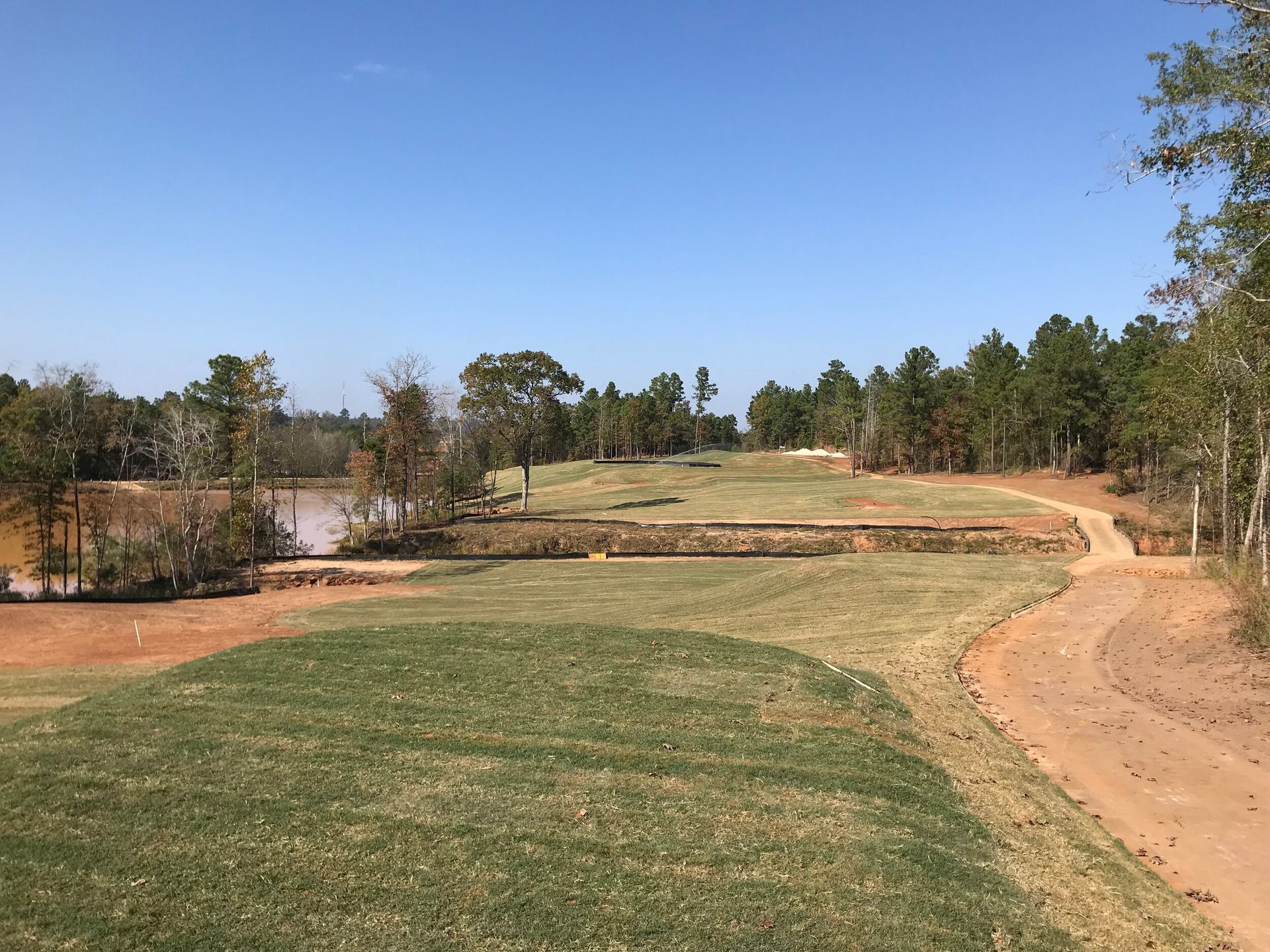 Curving dirt path beside a grassy field and pond under a clear blue sky