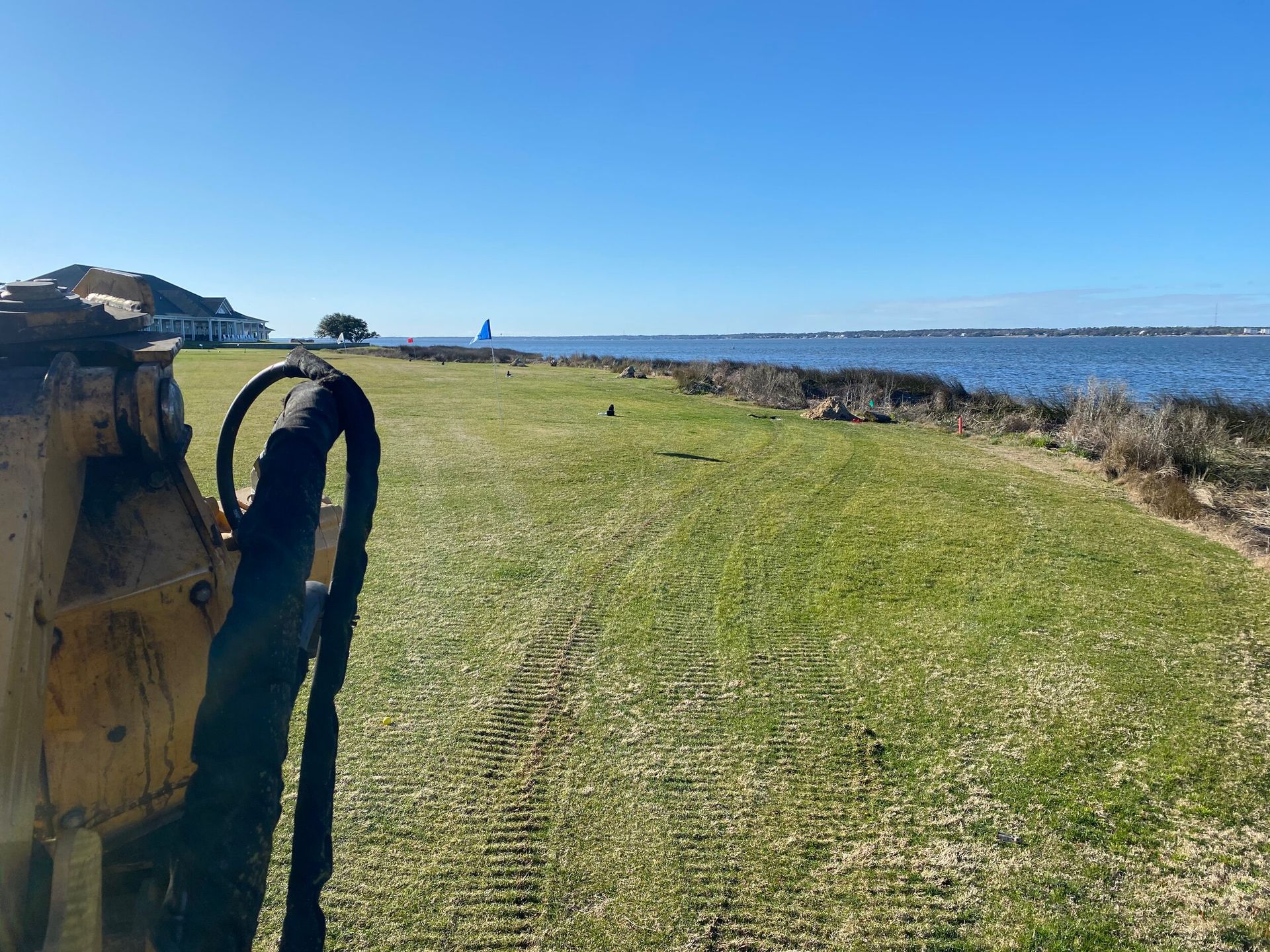 Grassy coastal path beside blue water under a clear sky, with a yellow machine on the left