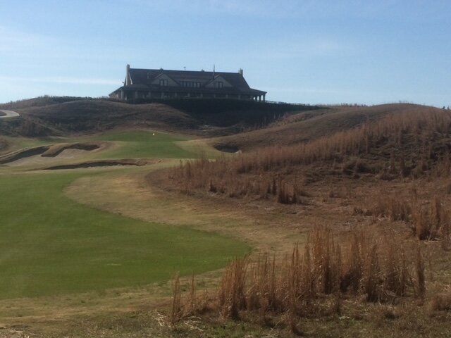 A large building on a hill overlooking a green golf course with sandy dunes under a blue sky