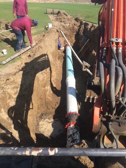 Workers installing a blue pipe in a deep trench beside a red excavator