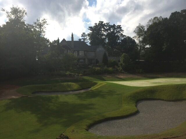 Golf course green and sand bunker with a clubhouse and trees in the background under cloudy sky