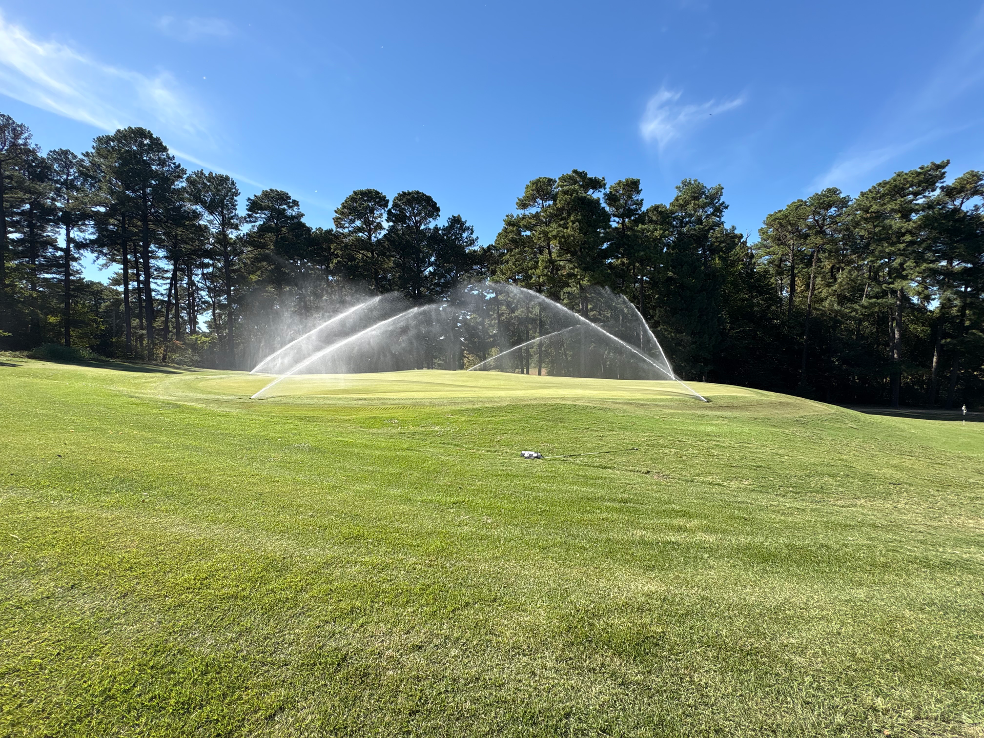 Sprinklers spraying water over a green field with trees under a clear blue sky