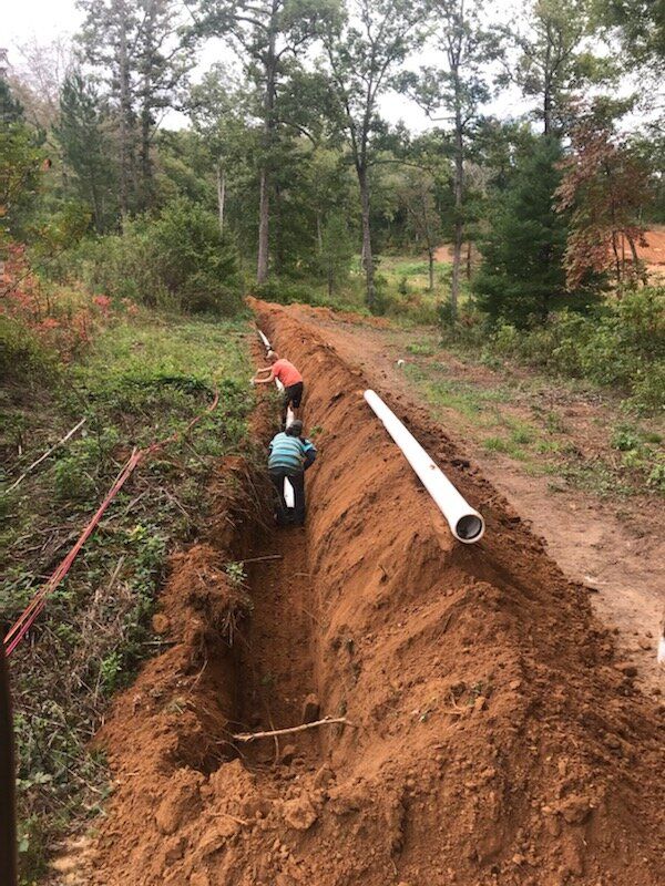 Workers lay a large white pipe in a dirt trench beside a wooded road.