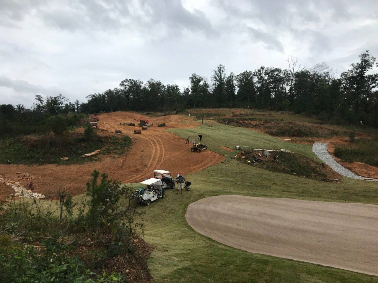 Dirt bike track with riders on a winding dirt trail under a cloudy sky, bordered by grassy hills and trees