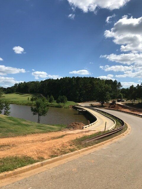 Curving paved road beside a small pond, grassy slopes, and a forest under a blue sky with scattered clouds