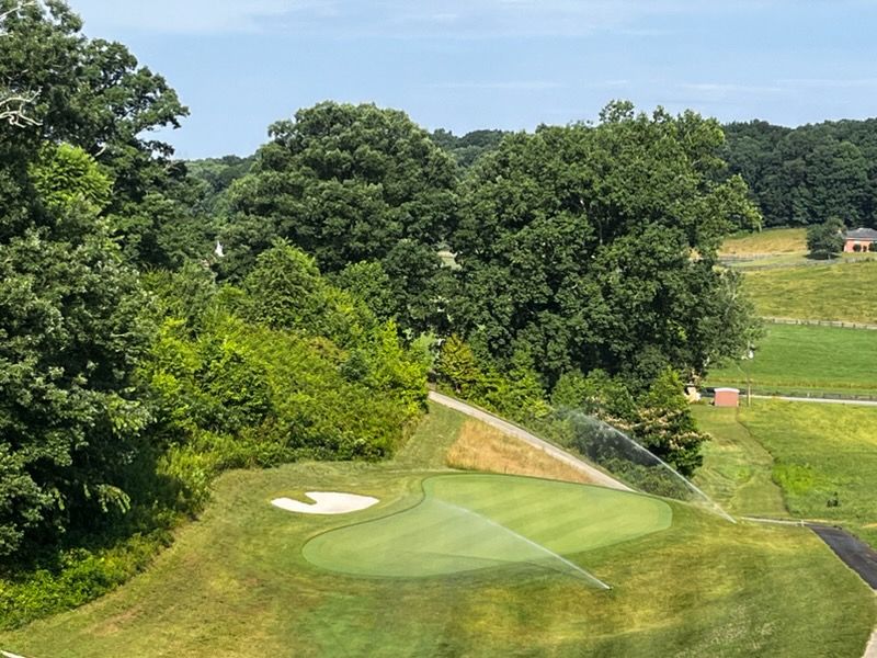 Golf course fairway with sand bunker, tree-lined hills, and a sprinkler watering the green
