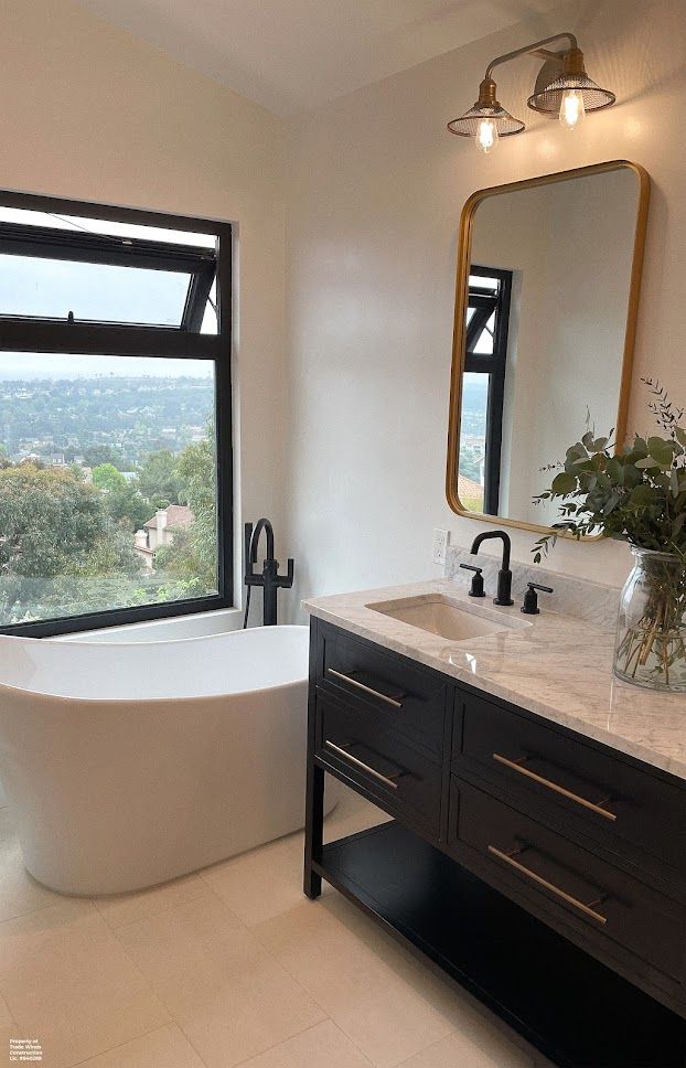 Modern bathroom with a view: a white tub, black vanity, gold-framed mirror, and large window overlooking a landscape.