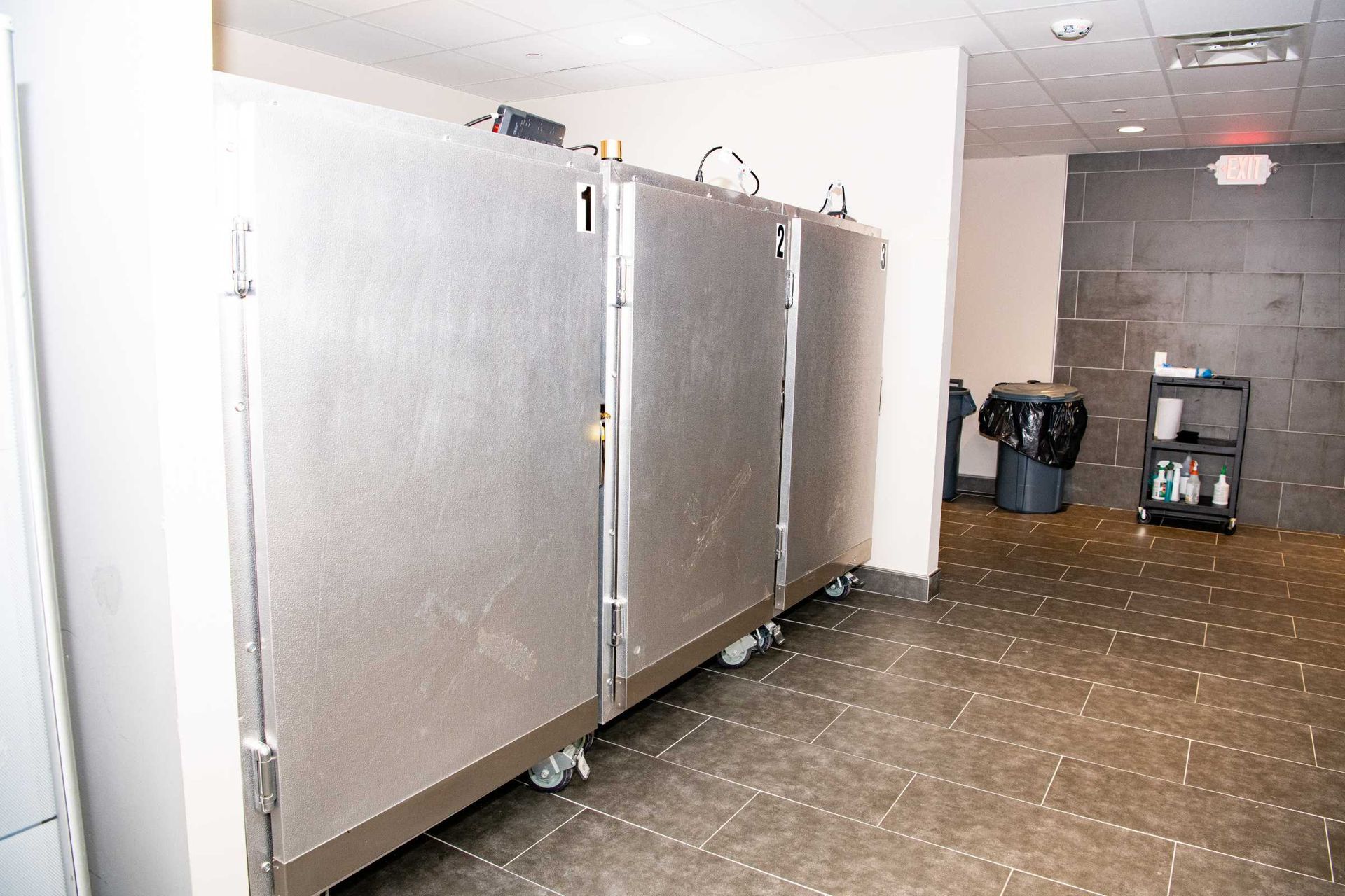 Row of stainless steel commercial refrigerators in a tiled kitchen or storage area