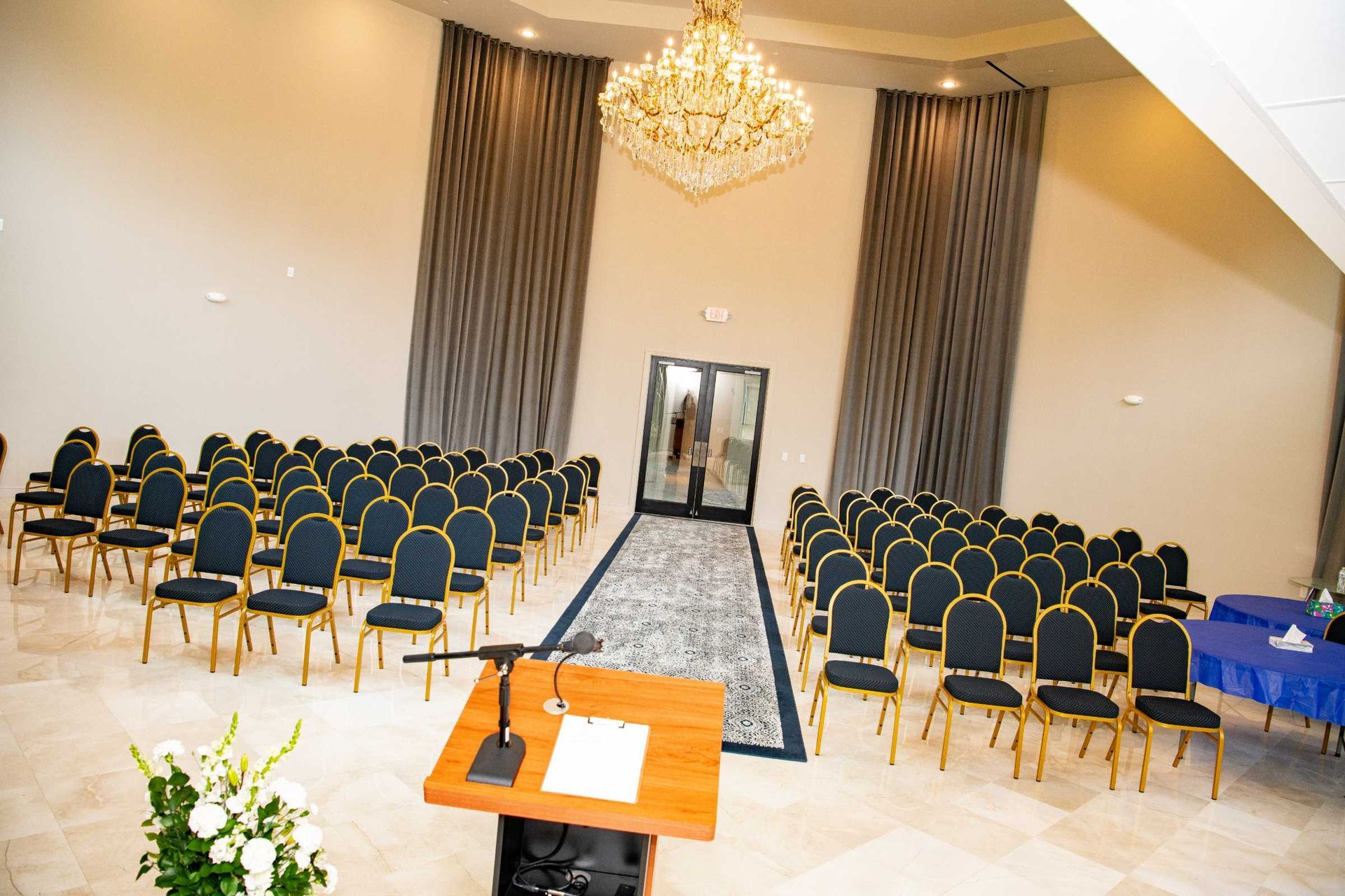 Conference hall with rows of blue-and-gold chairs, chandelier, and a central aisle leading to a doorway.