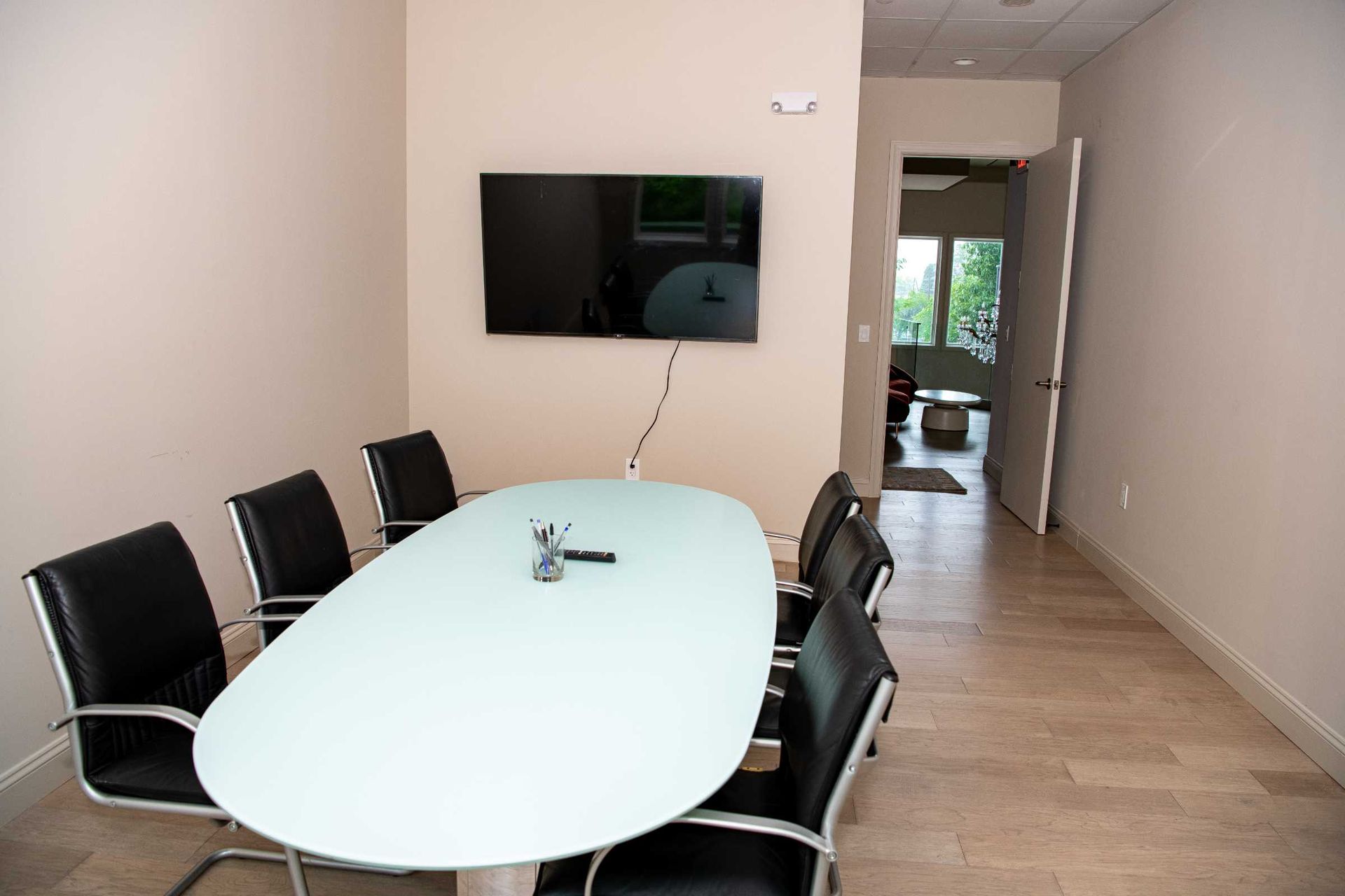 Modern conference room with oval white table, black chairs, wall-mounted TV, and hallway view
