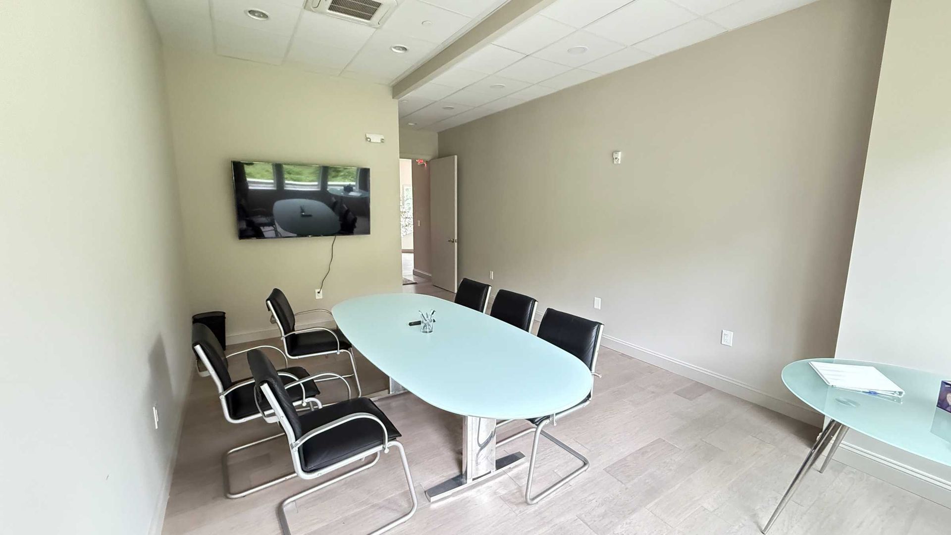 Empty conference room with oval table, black chairs, wall-mounted TV, and beige walls.
