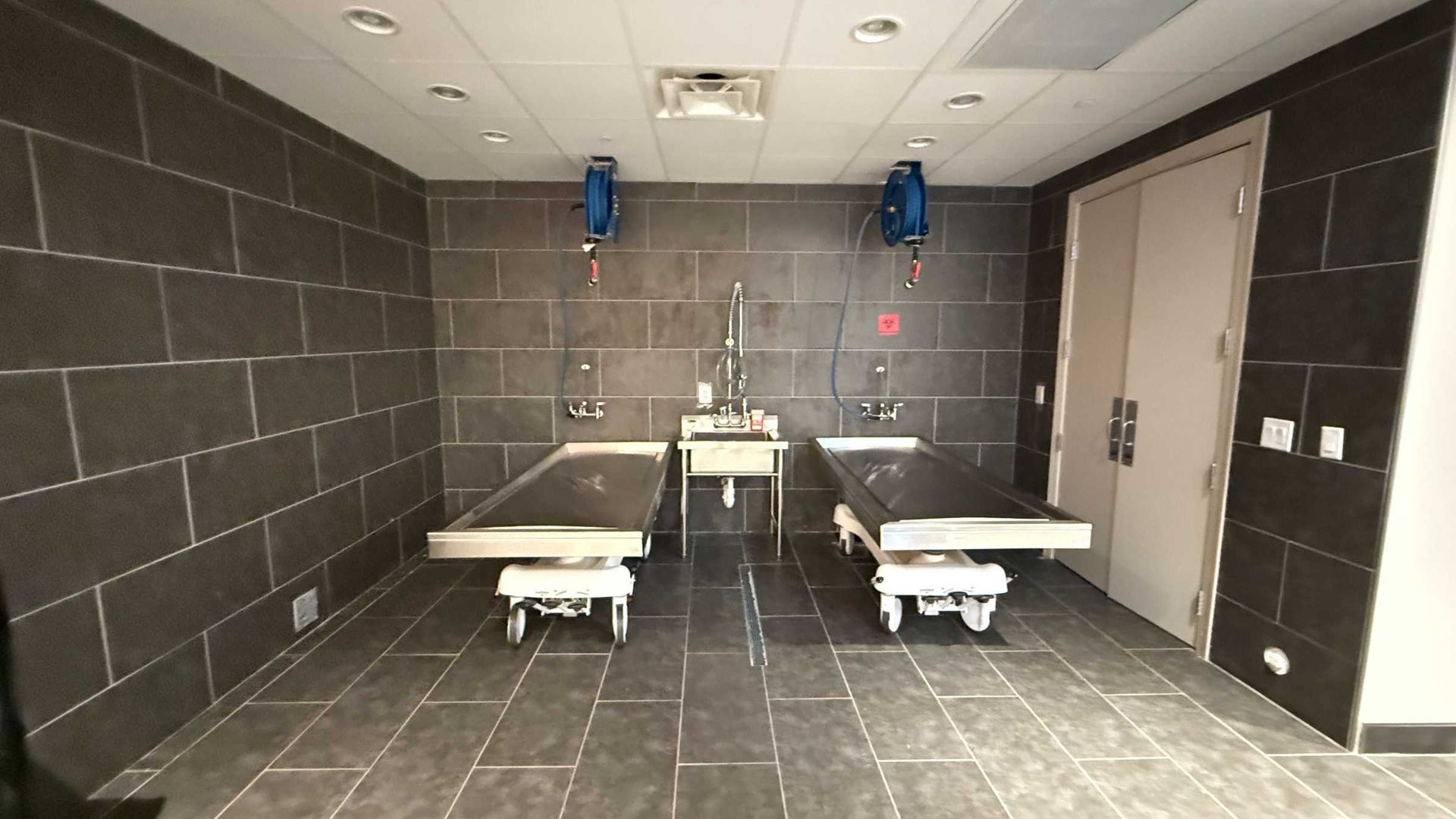 Clean restroom with two white sinks, wall-mounted dryers, and two toilet stalls in a gray tiled room.