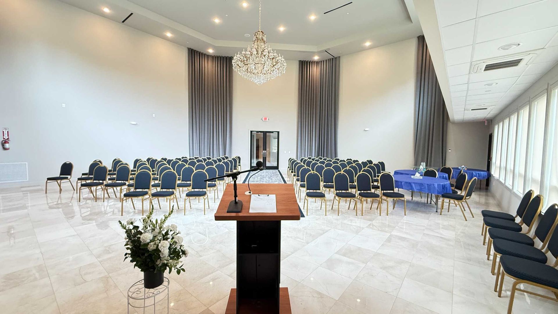 Bright conference hall with rows of chairs, a podium, chandeliers, and tall windows