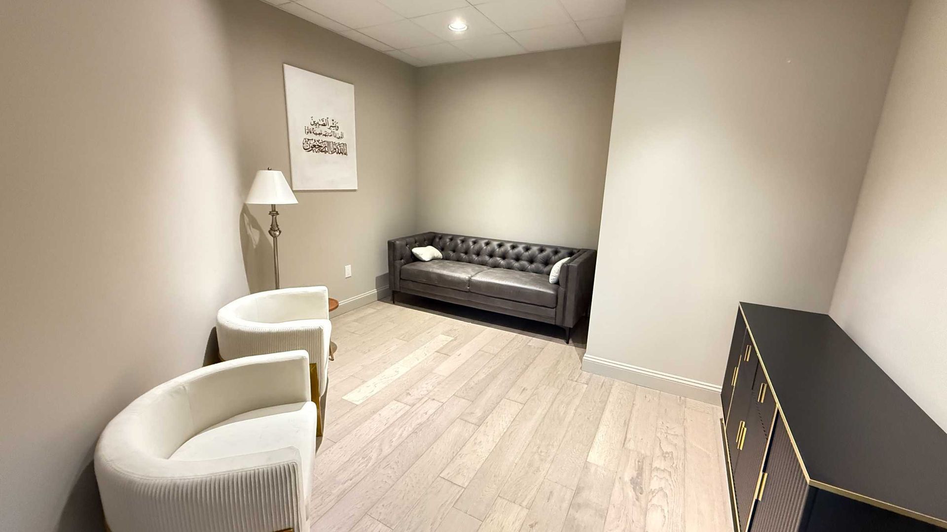 Small waiting room with beige walls, two white chairs, a black sofa, floor lamp, and a sideboard.