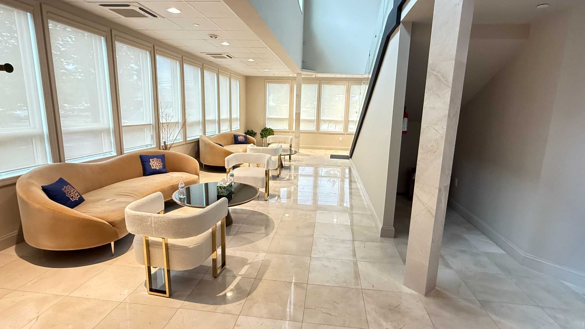 Bright modern lobby with beige seating, small tables, and large windows along a polished marble floor