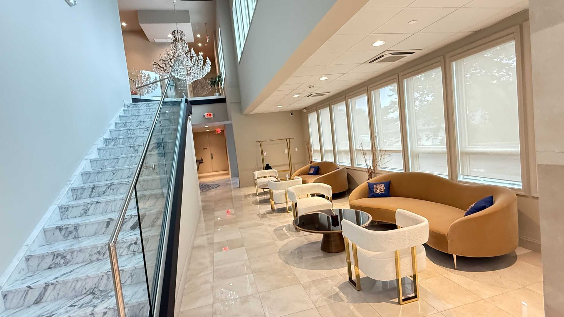 Bright modern lobby with staircase, chandelier, and tan seating by large windows