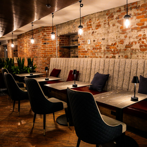 Cozy brick-walled restaurant with empty tables, black chairs, warm pendant lights, and wooden ceiling beams