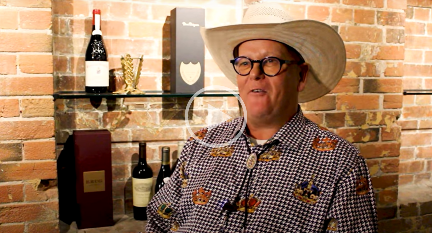 Man in a cowboy hat and glasses speaking before a brick wall with wine bottles and framed art
