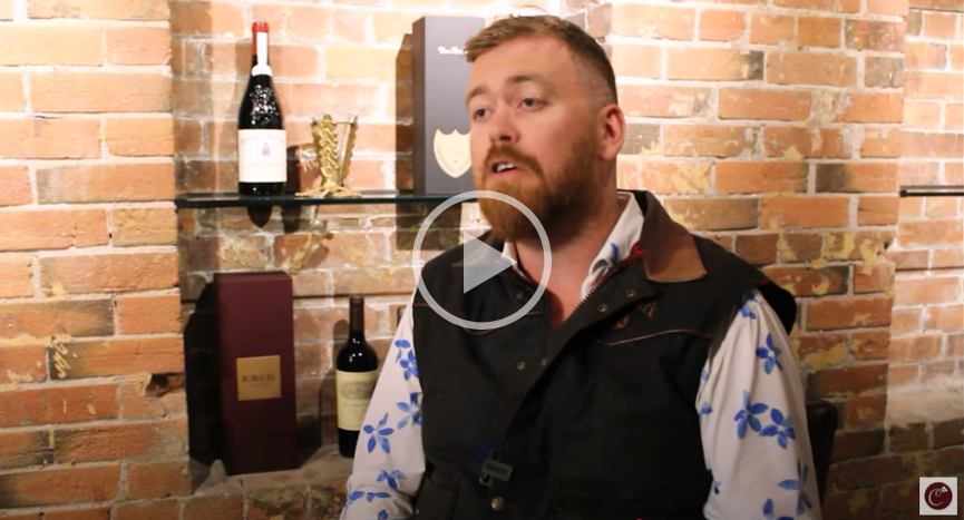 Man standing before a brick wall and wine shelf, speaking in a black vest and patterned shirt