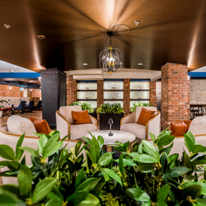 Stylish lounge with white chairs, orange pillows, brick columns, and green plants under warm lighting