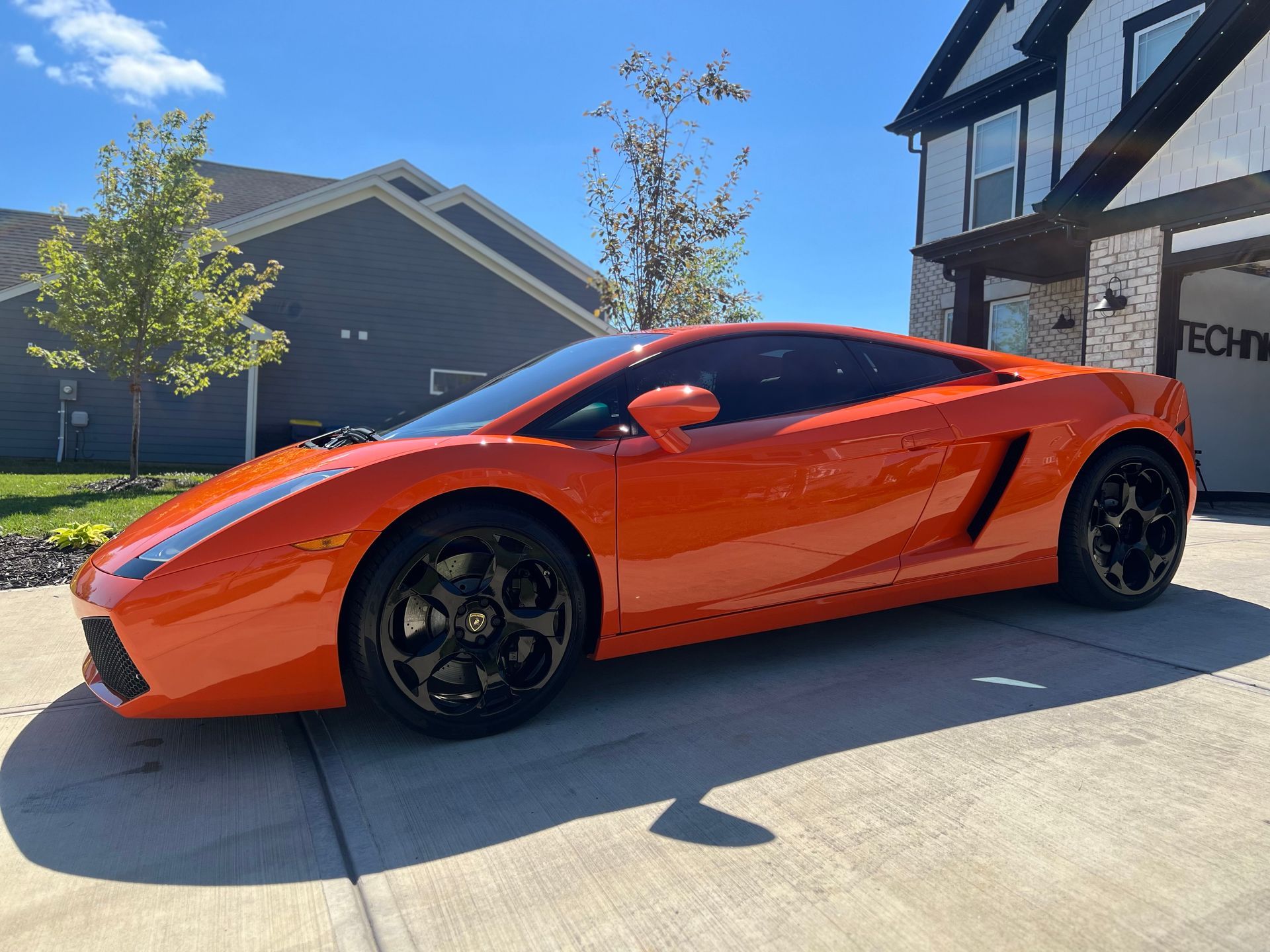 Bright orange Lamborghini parked in front of a modern house on a sunny day. Black wheels.