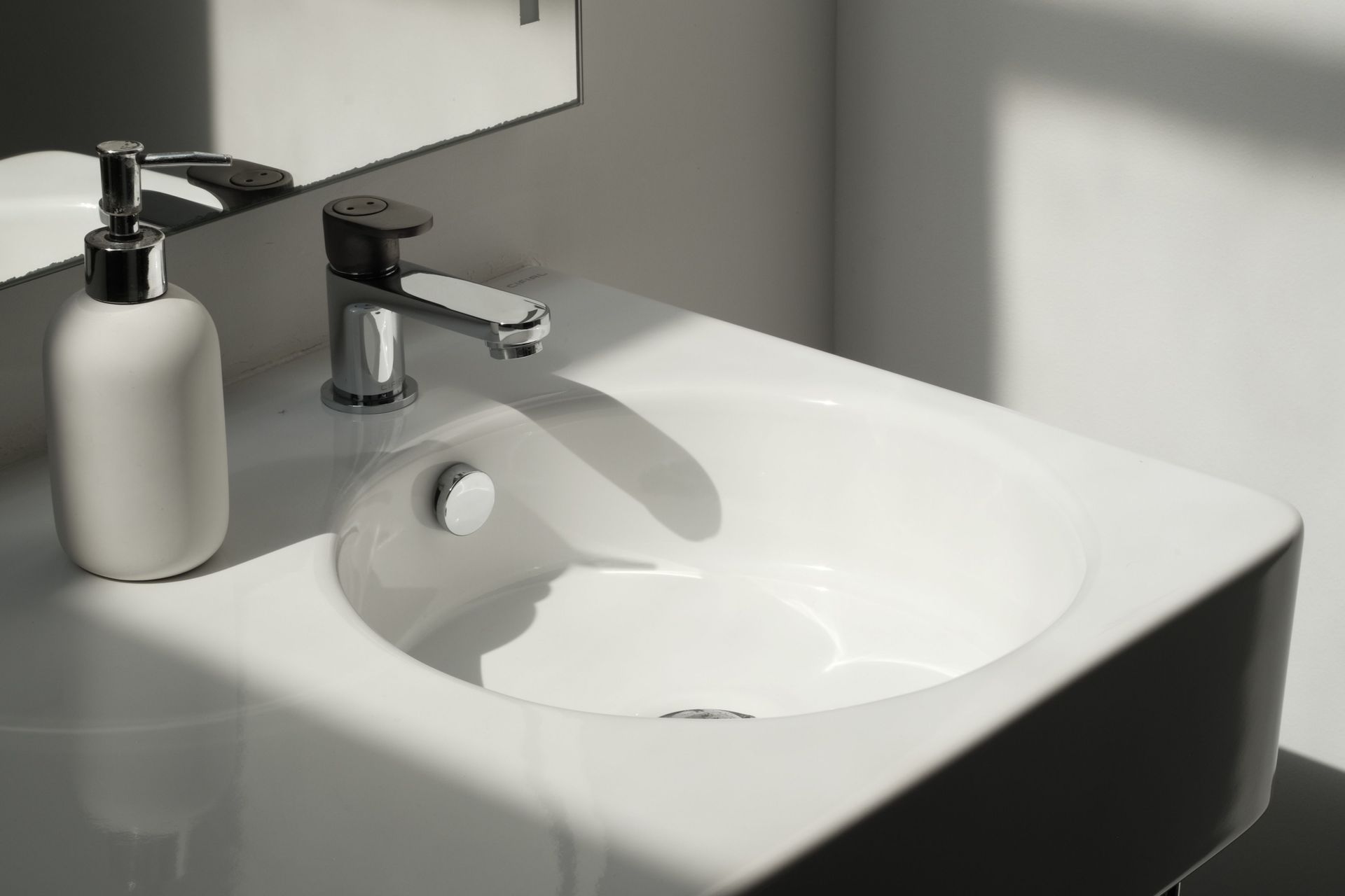 White bathroom sink with chrome faucet and soap dispenser on a countertop, lit by sunlight.