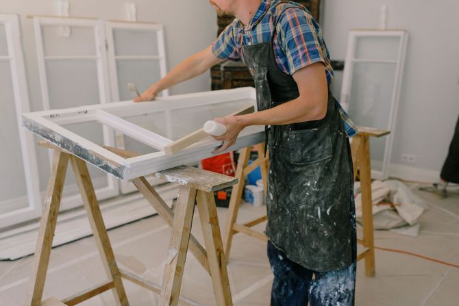 Person painting a white window frame in a workshop, with paint-covered clothes and tools nearby