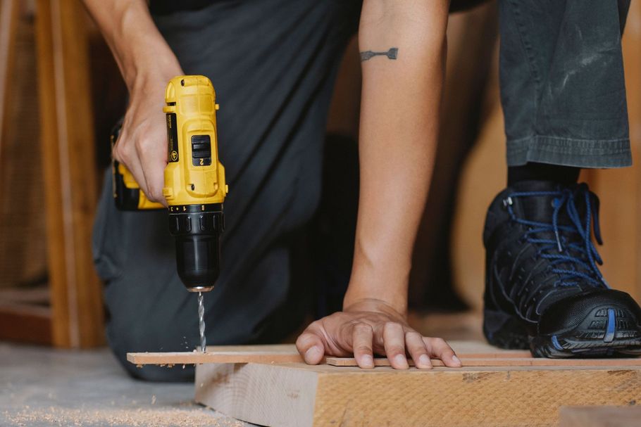 Person drilling into a wooden board while another steadies it on a floor indoors