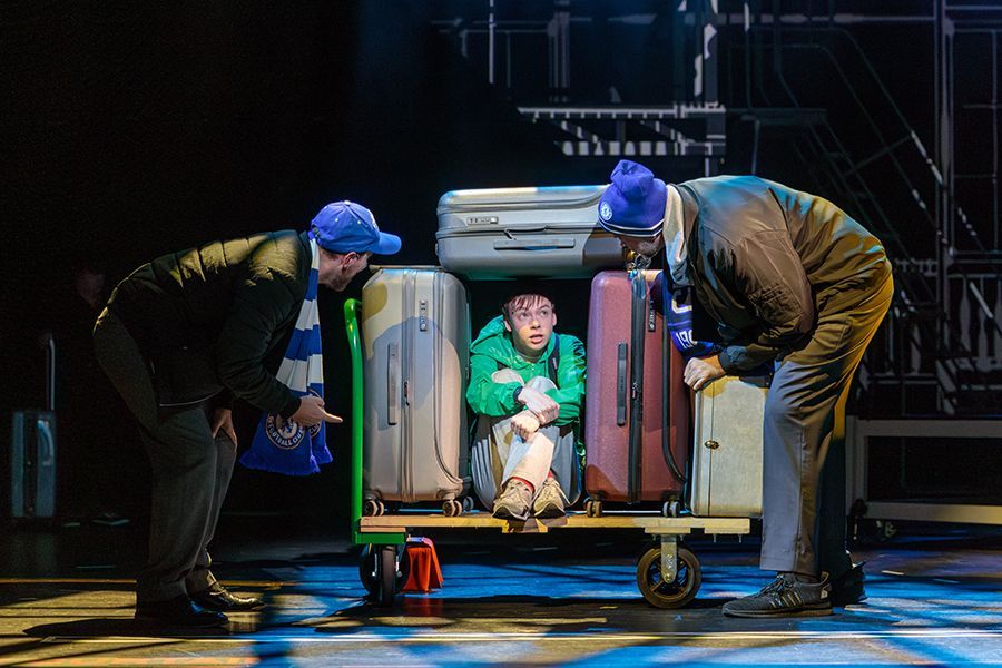 Two men in blue hats and scarves look at a person in green, surrounded by luggage on a cart.