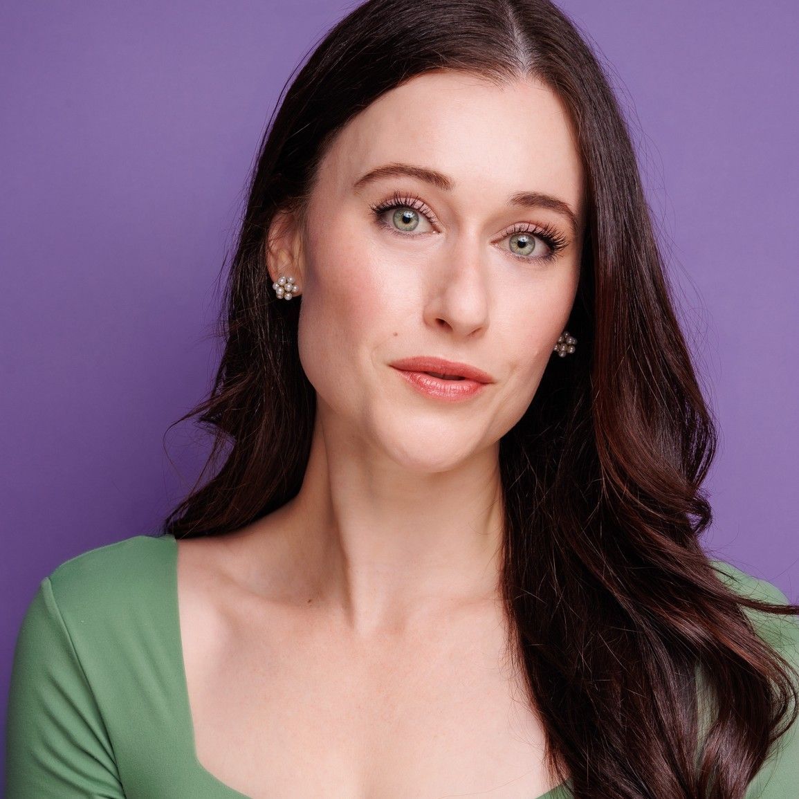 Portrait of a woman with long dark hair against a purple background, wearing a green top and earrings