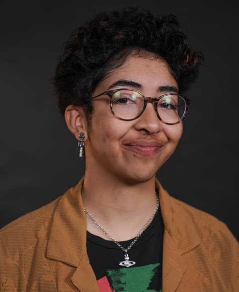 Studio portrait of a person with glasses, short curly hair, and a tan jacket, smiling against a dark background