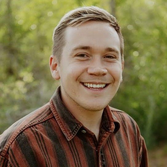 Smiling person in a red striped shirt outdoors with green foliage behind them