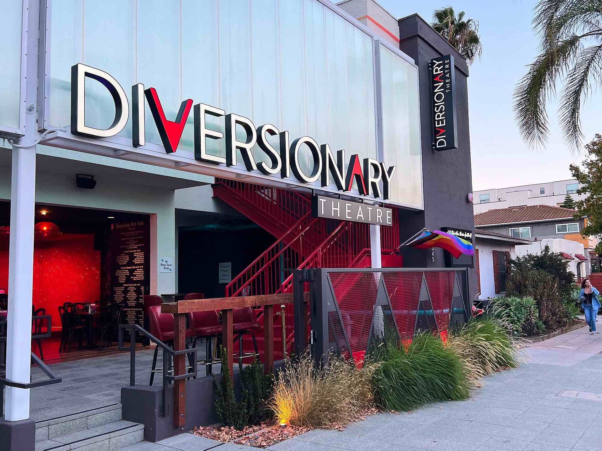 The Diversionary Theatre building features a red staircase, glass facade, and outdoor seating on a sidewalk.