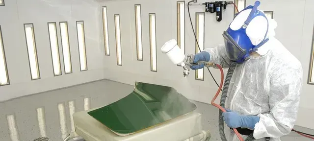 a man wearing a gas mask is spray painting a piece of metal in a paint booth .