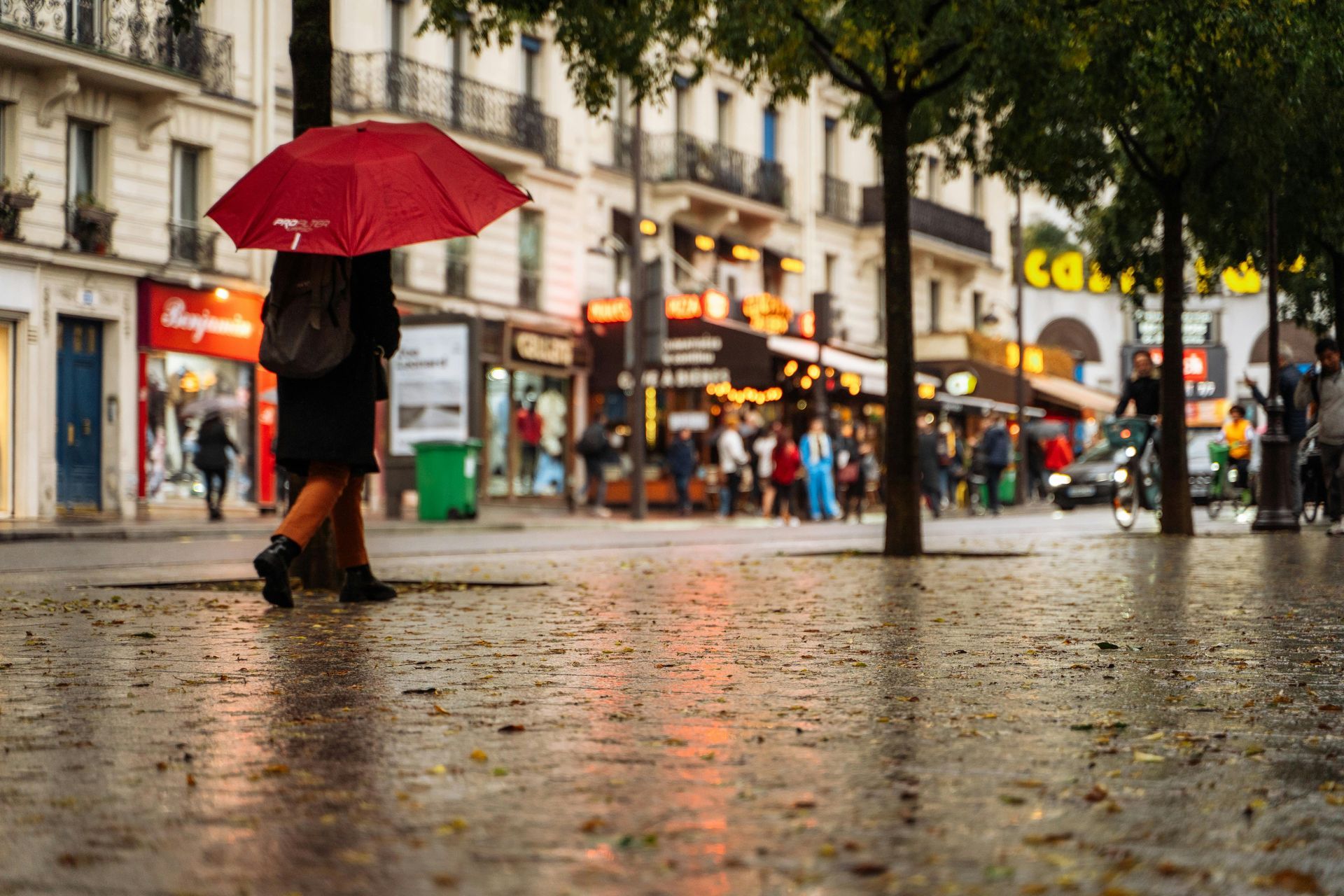 Une personne marche dans une rue mouillée, sous un parapluie rouge, avec des devantures de magasins
