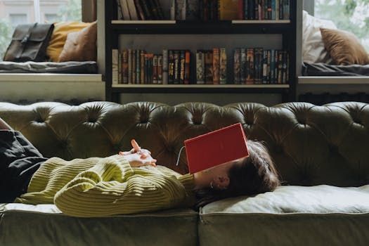 Une personne dort sur un canapé vert, un livre rouge sur le visage, avec des étagères en arrière-plan. Pièce ensoleillée.