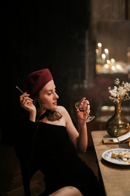 Femme en béret et robe noire, tenant un verre de vin et une cigarette à une table éclairée à la bougie.