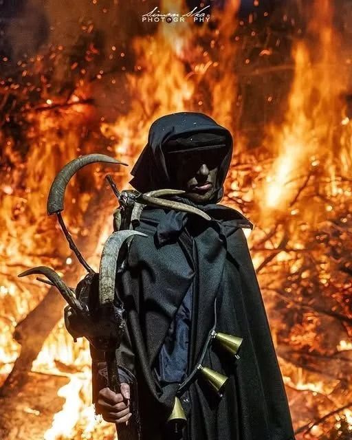 Una persona con un mantello nero e cappuccio è in piedi davanti a un falò, tenendo in mano un bastone con campanelli e lame ricurve.