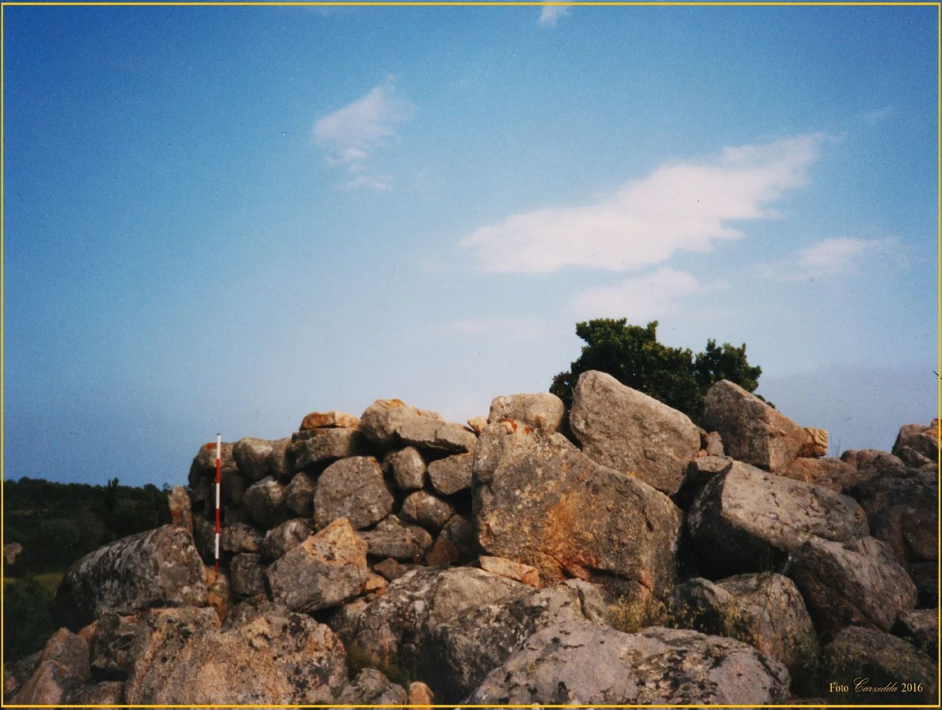 Un muro di pietra sotto un cielo azzurro, forse rovine. Un albero è in cima al muro.