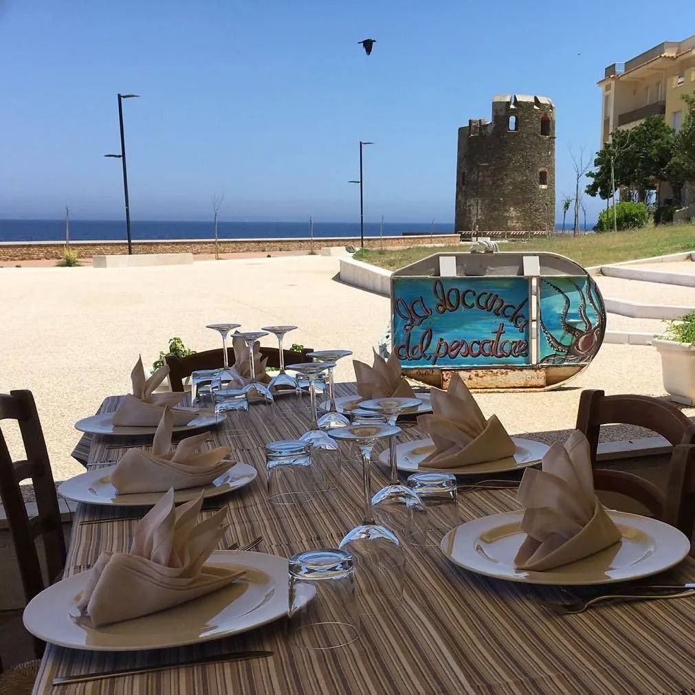 Tavolo da ristorante all'aperto apparecchiato per cenare, sullo sfondo l'oceano e una torre di pietra.