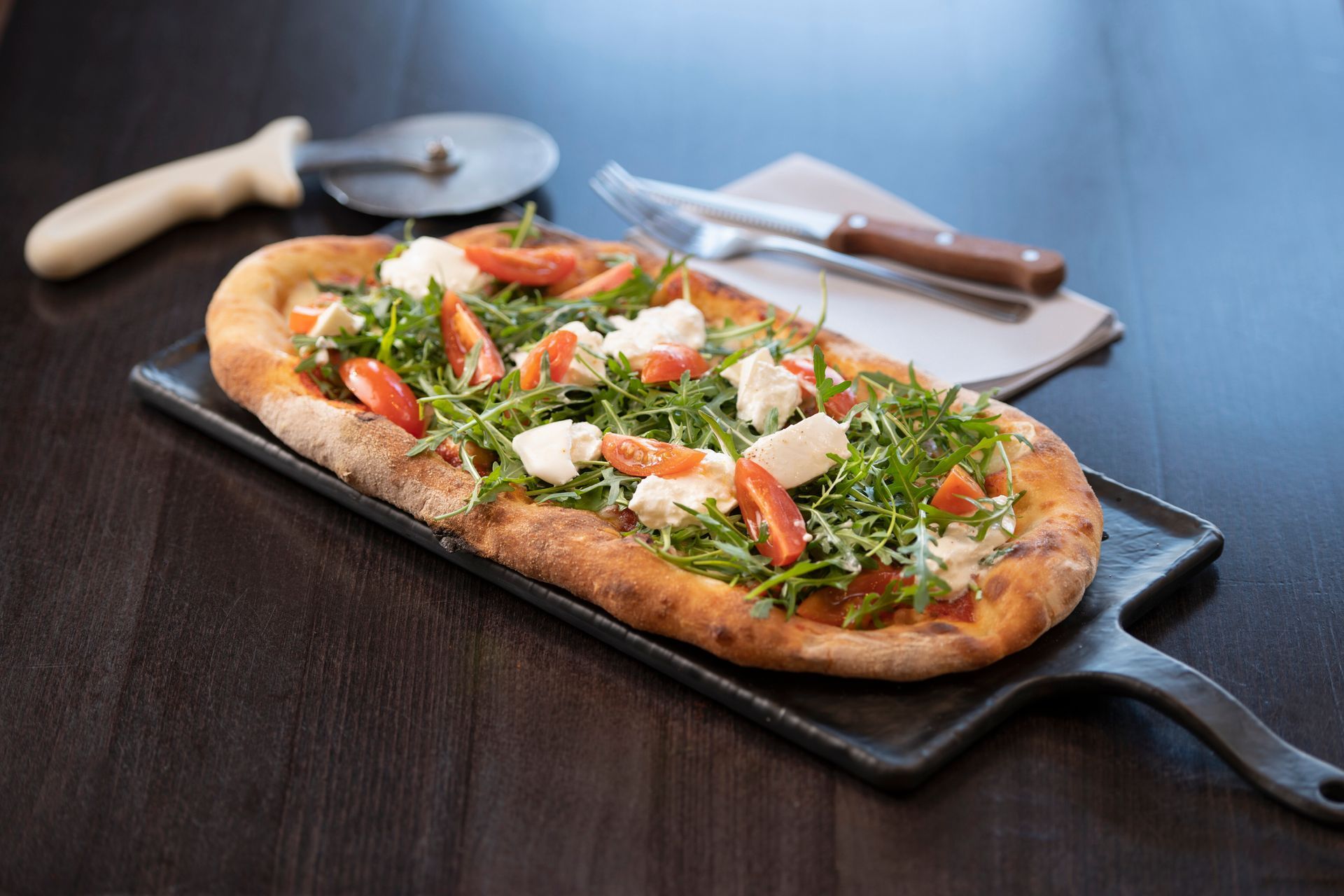 Pizza piatta condita con rucola, pomodori e formaggio su un piatto da portata nero con rotella tagliapizza e posate.