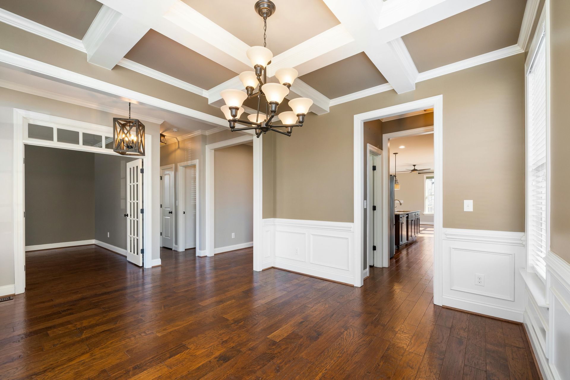 A bright room with wood floors, beige walls, white wainscoting, a coffered ceiling, and a traditional chandelier.
