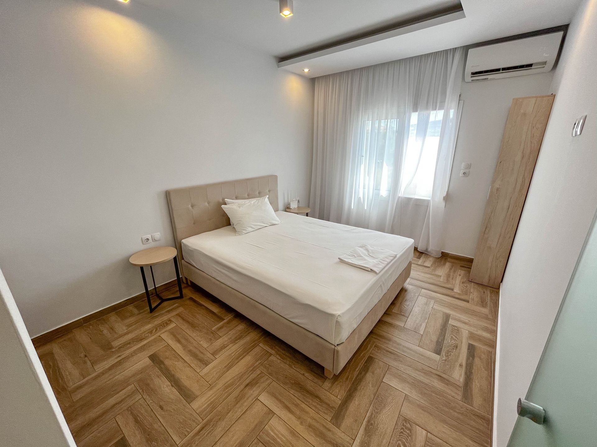 Bedroom with a bed, small table, and closet. Features wooden floors and a window with white curtains.