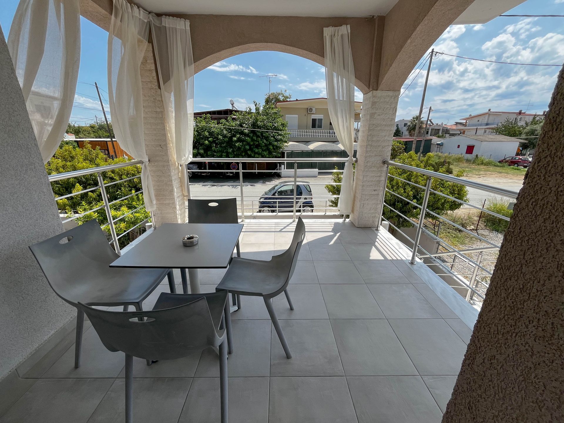 Balcony with table and chairs overlooking a street. White curtains, beige columns, and a sunny sky.