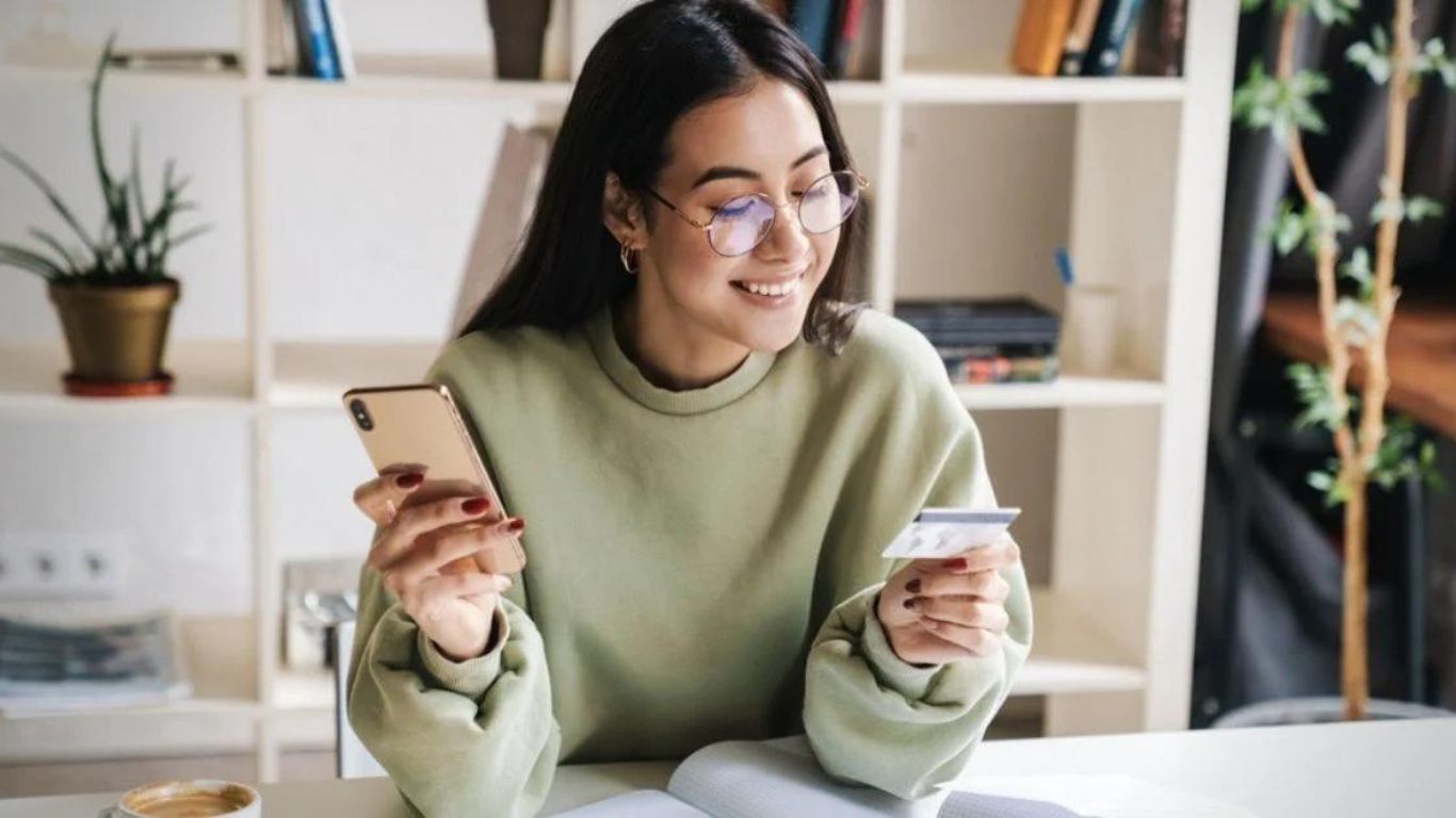 A woman is sitting at a table holding a credit card and a cell phone.