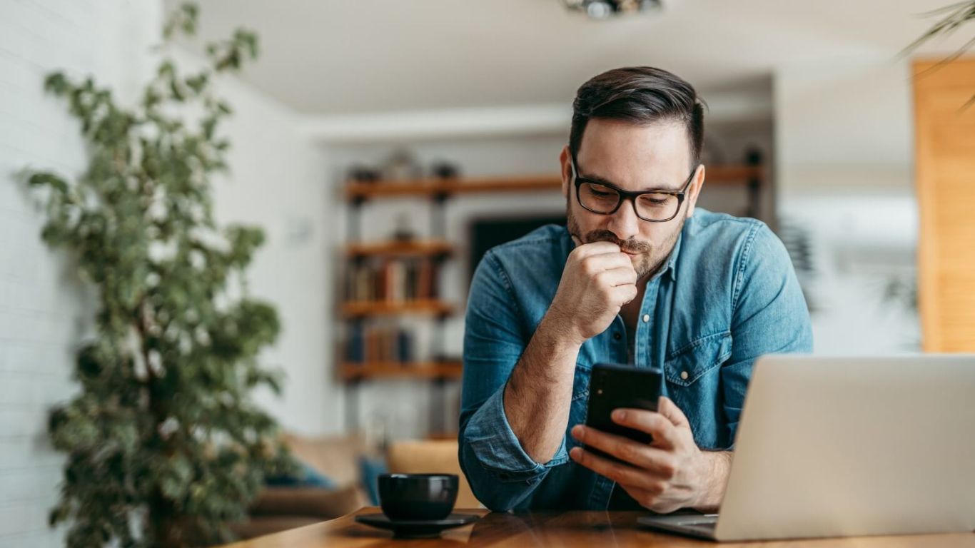 A man is sitting at a table using a laptop and a cell phone.