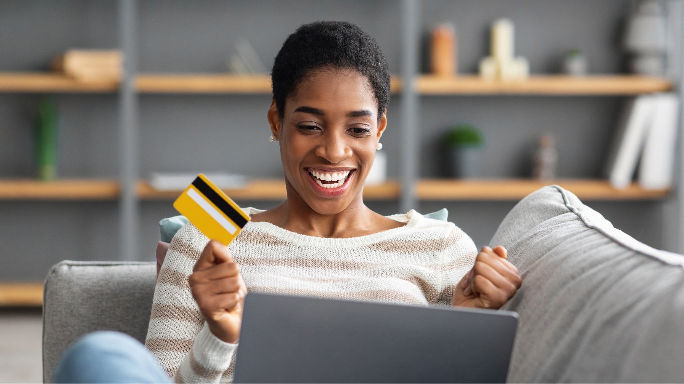A woman is sitting on a couch holding a credit card and a laptop.