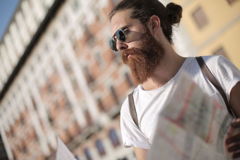 Hombre con gafas de sol y barba, vestido con una camiseta blanca, de pie al aire libre con un edificio borroso detrás de él.