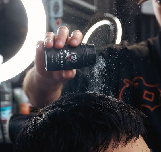 Una mano espolvorea polvos sobre cabello oscuro durante un corte de pelo, con un brillante aro de luz de fondo.