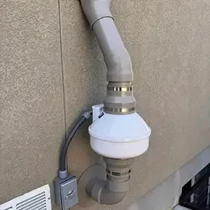 Close-up of an exterior radon fan installation: sealed pipe couplings, conduit routed flat against the wall, and a service disconnect switch within reach